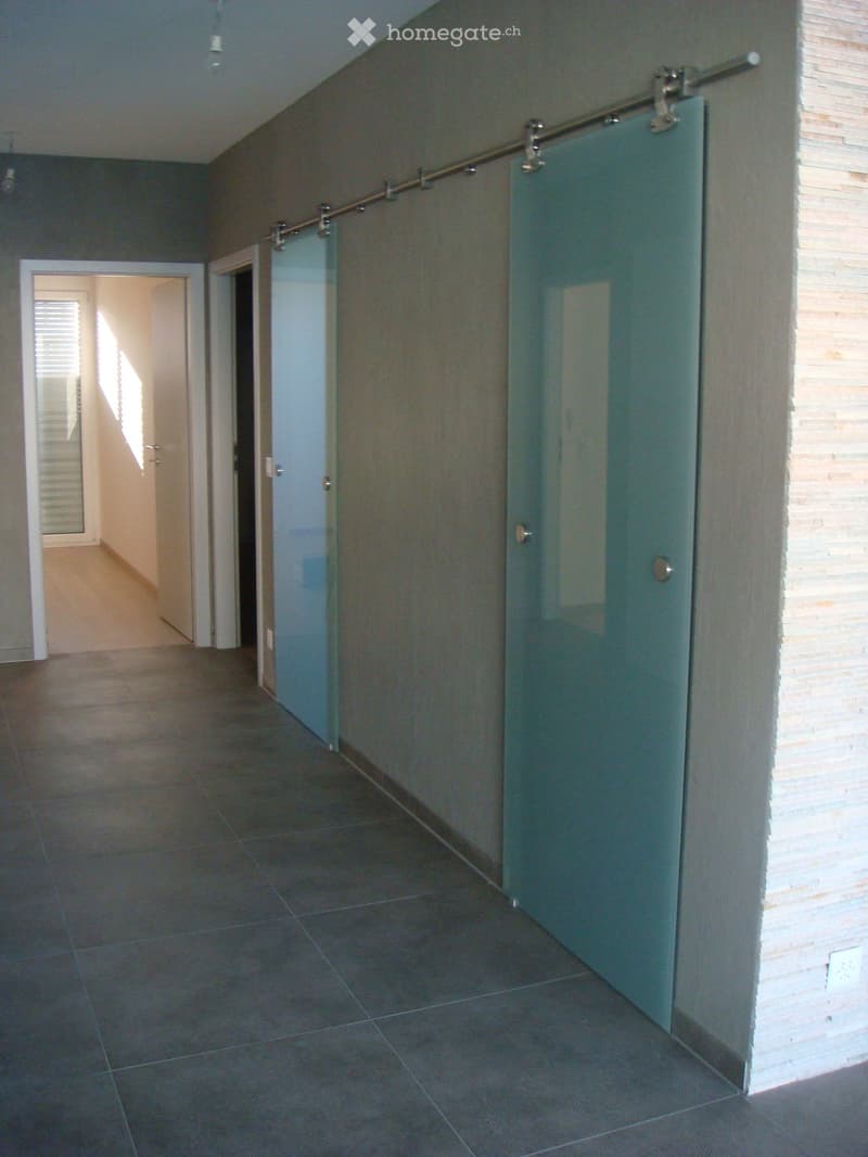 Modern hallway with dark tiled flooring, grey walls, and glass sliding doors on one side. The hallway features a sleek, contemporary design with a visible open doorway leading to another room.