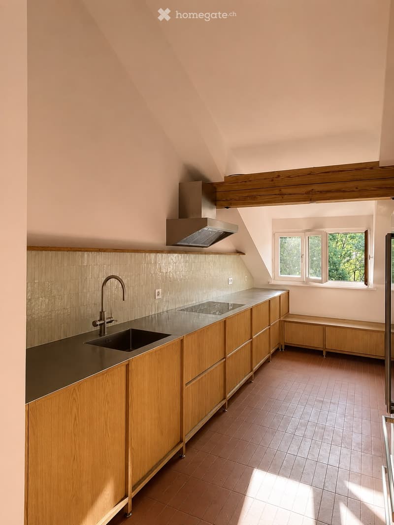 Modern kitchen with wooden cabinets, stainless steel sink, and range hood. The space features a slanted ceiling and large windows allowing natural light to fill the room.