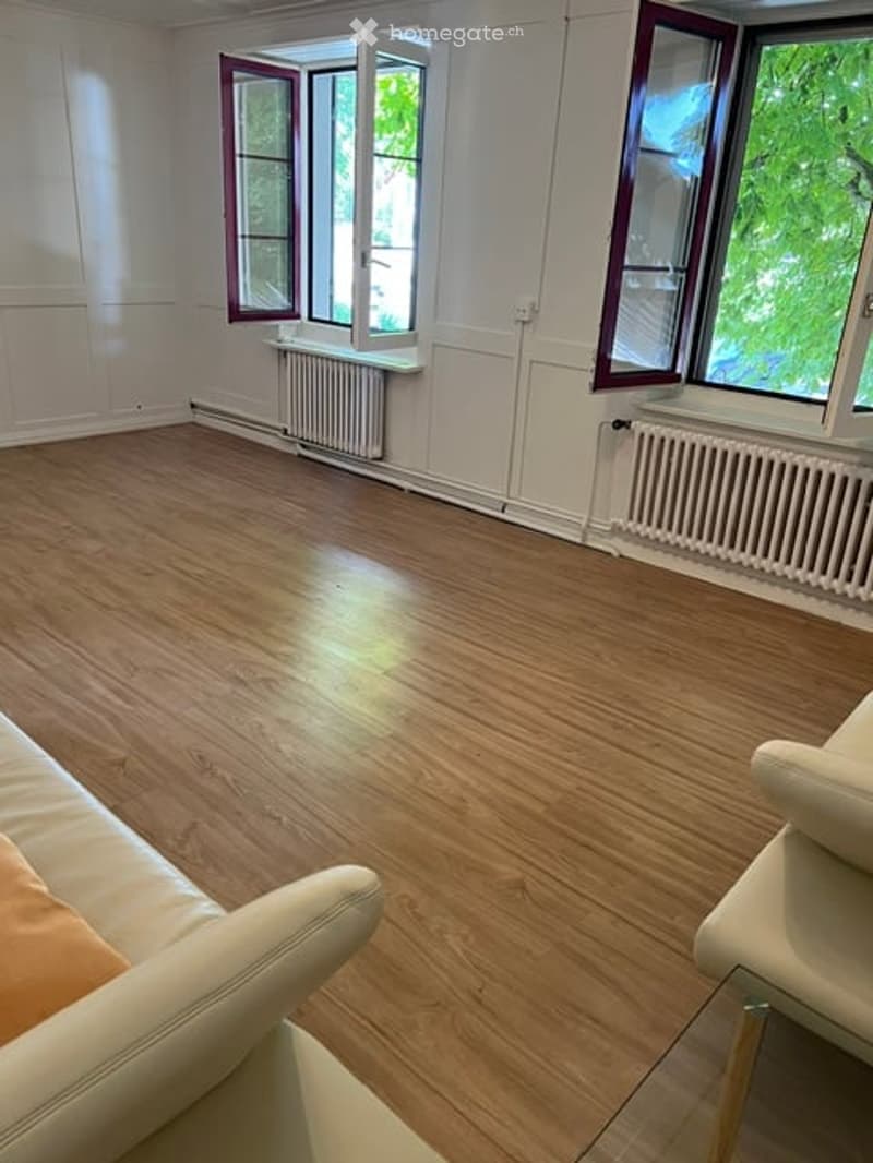 Bright, unfurnished room with wooden flooring, white walls, large windows with red frames, radiators, and a small sofa and table in the corner.