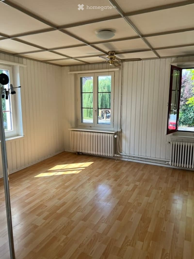 Empty room with wooden flooring, white paneled walls, and large windows allowing natural light. Radiators are visible under the windows, and a ceiling fan is mounted on the ceiling.