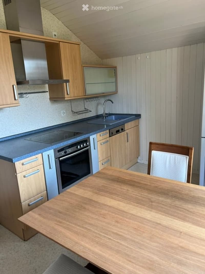 Modern kitchen with light wooden cabinets, stainless steel appliances, and a central dining table with chairs.