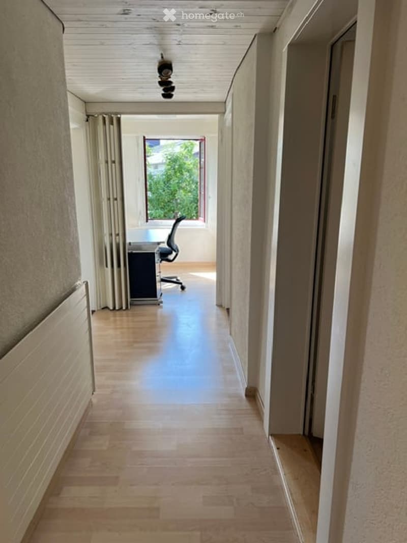 A narrow hallway with wooden flooring, white walls, and a window at the end. There is a radiator on the left wall and a small office area visible at the end of the hallway.