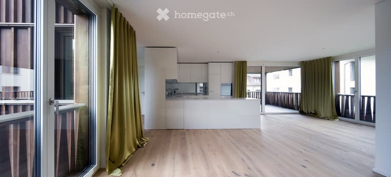 Open-plan living and kitchen area with light wooden flooring, white cabinetry, and large windows leading to a balcony.