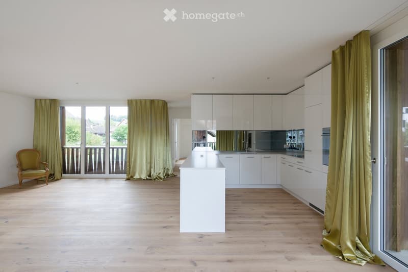 Open-plan kitchen and living area with light wooden flooring, white kitchen cabinets, a central island, and a green curtained window leading to a balcony.