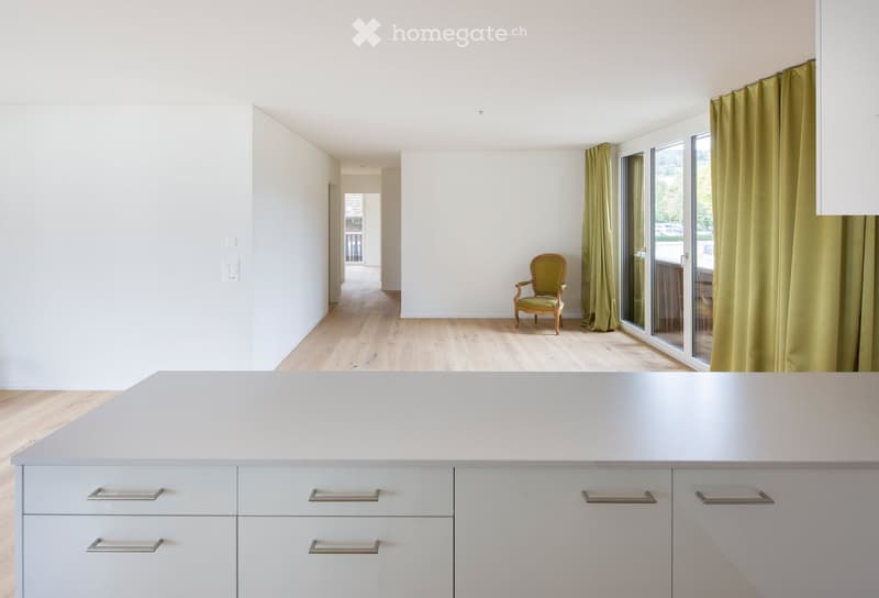 Bright, minimalist kitchen with light grey cabinets and a central island, featuring a single green armchair near large windows with green curtains.