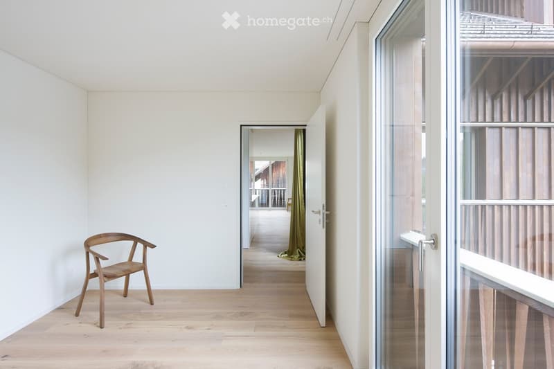 Bright hallway with light wooden flooring, white walls, and a single wooden chair near the wall. An open doorway leads to another room with a visible green curtain and a glimpse of a balcony area.