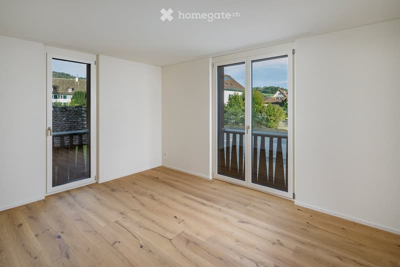 Empty room with light wooden flooring and white walls, featuring large sliding glass doors that open to a balcony with a view of the surrounding area.