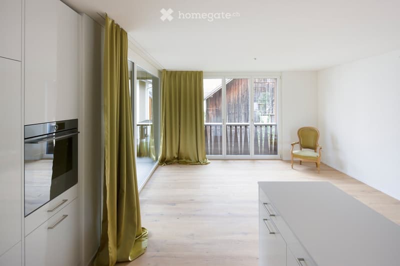Bright, modern room with light wooden flooring, white cabinets, a built-in oven, a single elegant chair, and large glass doors leading to a balcony.