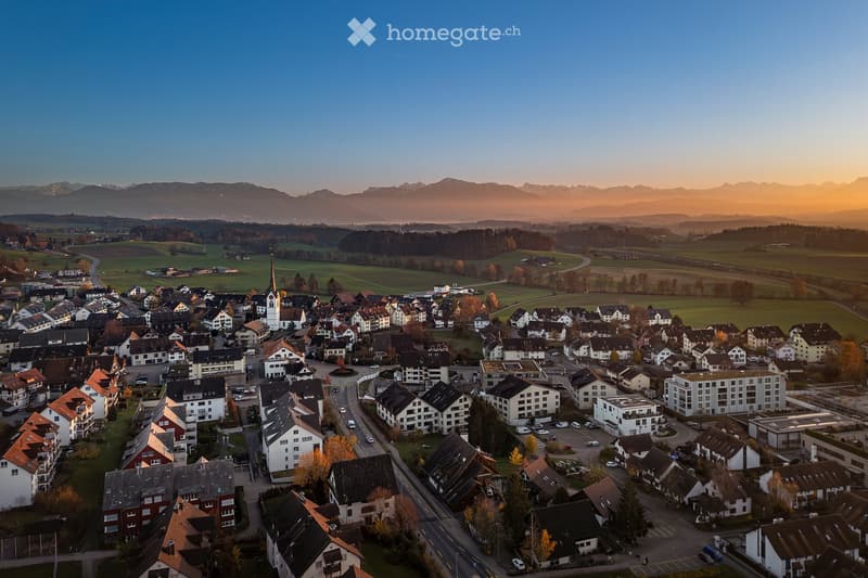 Aerial view of a small town nestled in a valley with surrounding mountains, featuring houses, roads, and a church spire.
