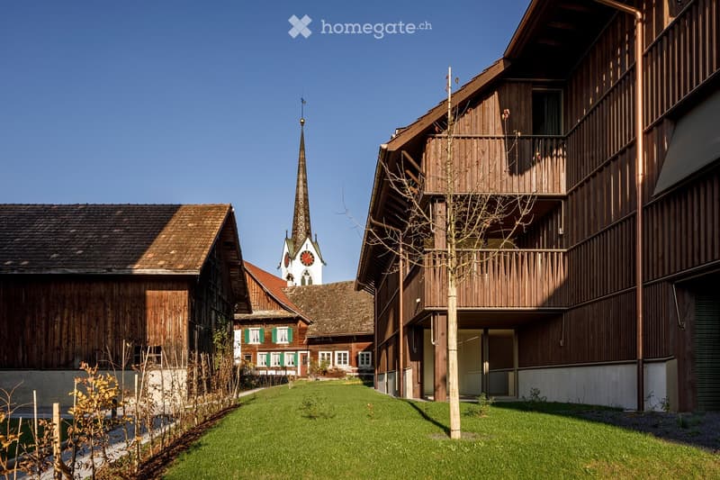 Traditional wooden houses with green shutters, a church tower with a clock, and a well-maintained lawn in a village setting.
