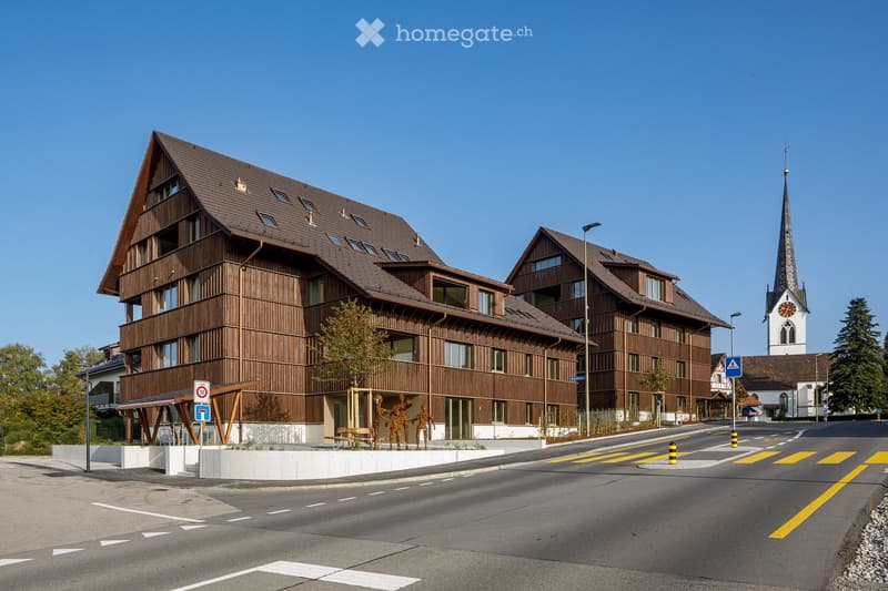 Modern multi-story wooden building with large windows and balconies, situated on a street corner with a church tower visible in the background.