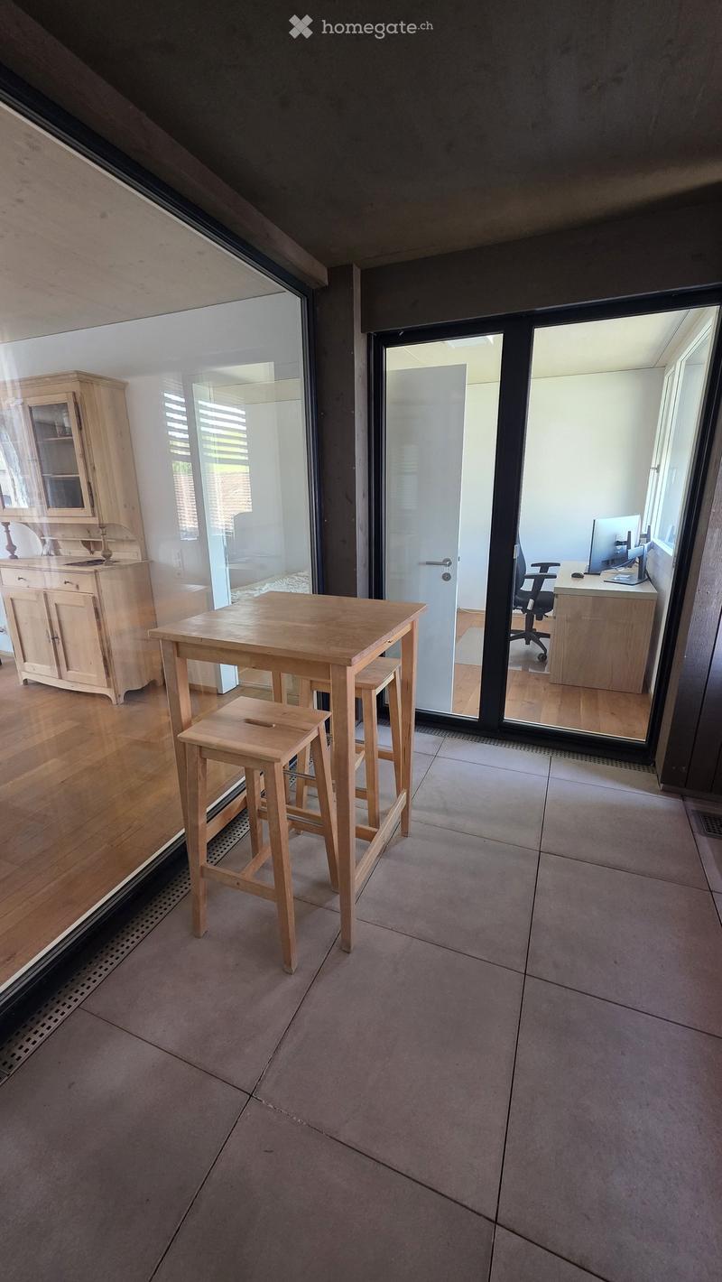A modern, well-lit room with a wooden table and stools, a wooden cabinet, and a desk with a computer in the adjacent space visible through large glass doors.