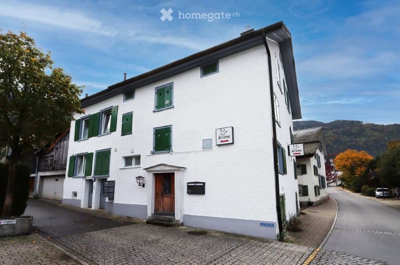 A two-story white building with green shutters and a wooden front door, situated on a cobblestone street with a sign reading "Krone" above the entrance.