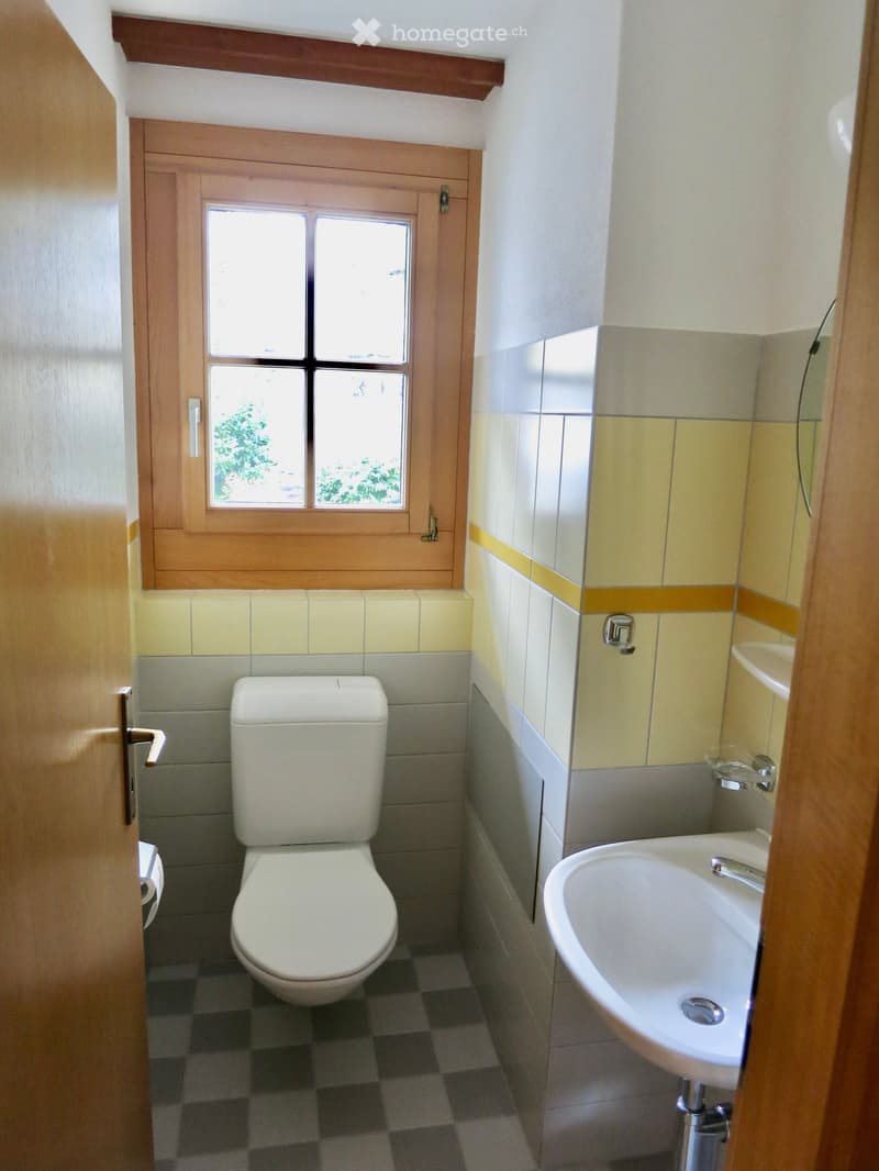 Small bathroom with a toilet, sink, and window. The walls are tiled in a two-tone design with yellow and gray tiles, and the floor has a checkered pattern.