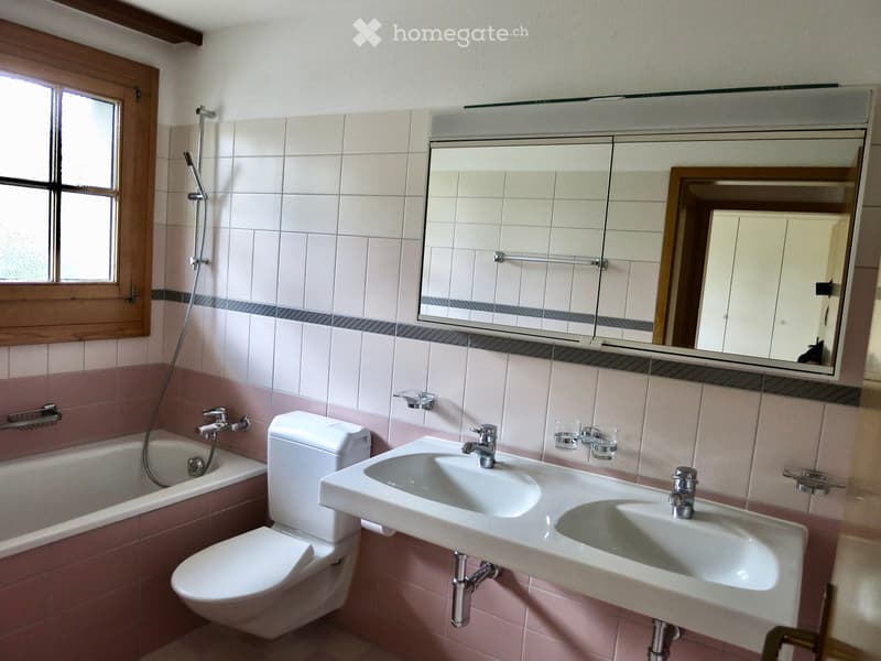 Bathroom with pink and white tiles, a bathtub, toilet, double sink, mirror, and a window with wooden frame.