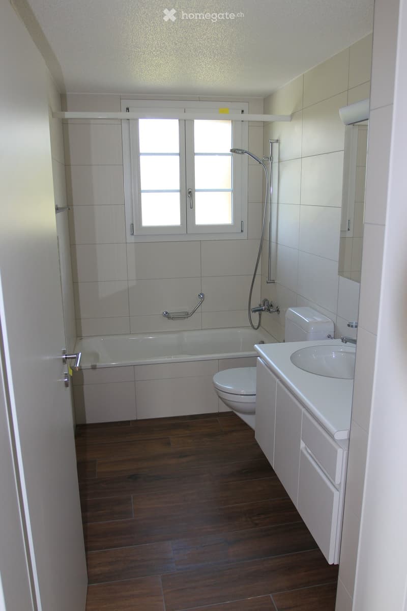 Modern bathroom with wooden flooring, white tiled walls, bathtub, toilet, sink, and showerhead.
