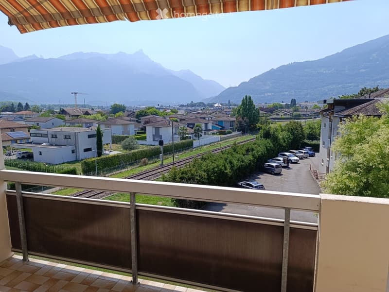 Balcony with a view of a town, railway tracks, and distant mountains. The balcony has a metal railing and tiled flooring.