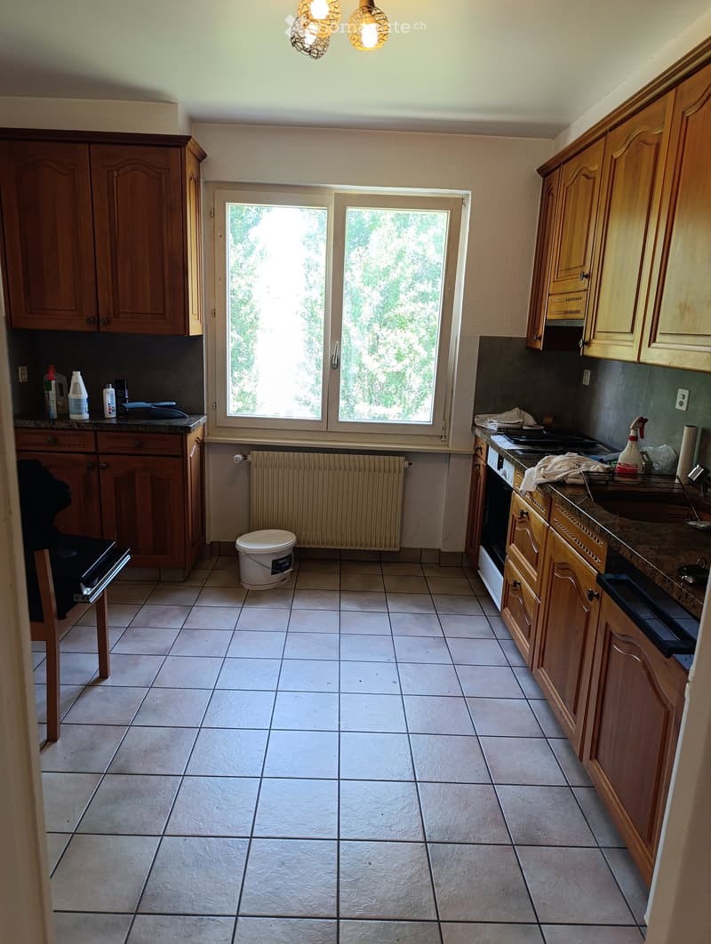 Kitchen with wooden cabinets, tiled floor, and a window with double doors leading outside. Cleaning supplies are on the counter, and a small stool is near the oven.