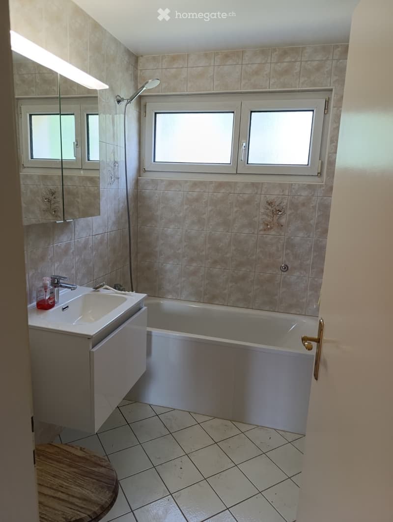 Modern bathroom with beige tiled walls, a white bathtub, a sink with a faucet, a mirror, and a showerhead. The floor is tiled with light-colored tiles.