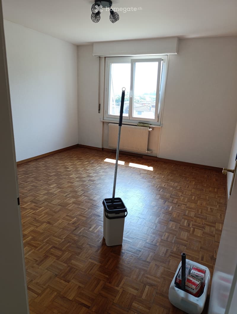 Empty room with wooden herringbone flooring, white walls, and a large window with a radiator below. Cleaning supplies and a trash bin are visible.