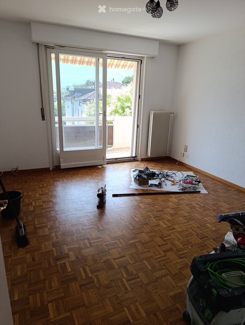Empty room with wooden parquet flooring, white walls, large sliding glass door leading to a balcony, and a radiator on the wall.