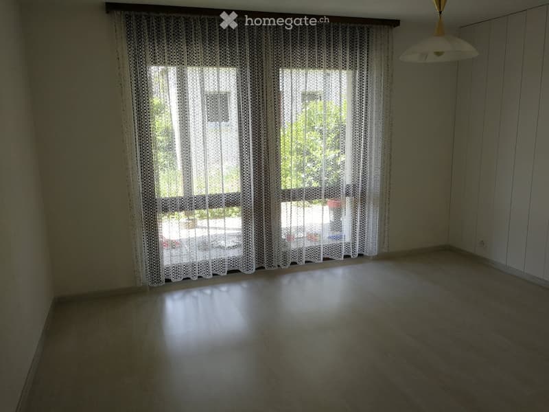 Empty room with wooden flooring, large windows covered with white lace curtains, and a hanging light fixture.
