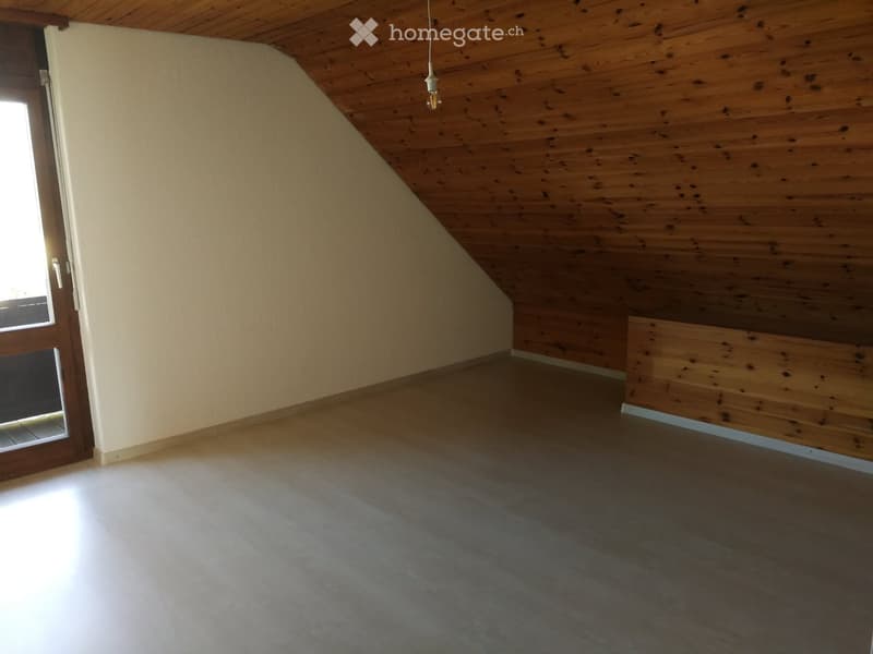 Empty attic room with wooden slanted ceiling, wooden floor, and a single door leading outside.