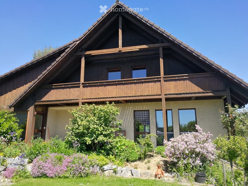 Two-story wooden house with a balcony, surrounded by a well-maintained garden with flowers and shrubs.