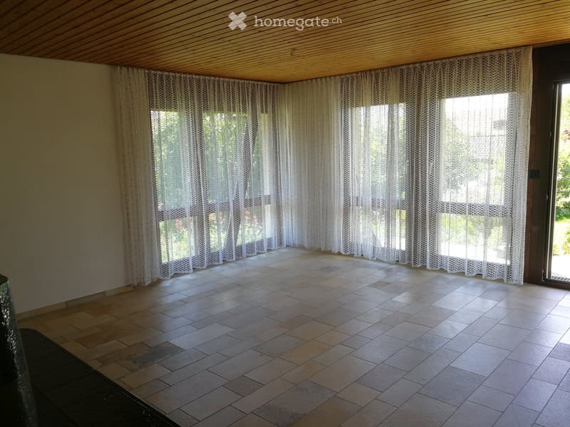 Empty room with tiled floor, large windows covered by sheer curtains, and a wooden ceiling.