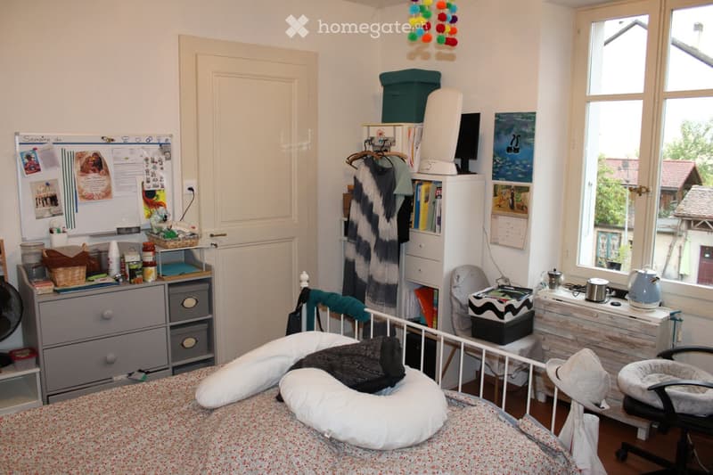 A cozy bedroom with a bed, a chair, a fan, a small table with various items, a refrigerator, a screen, and a cat bed near the window.
