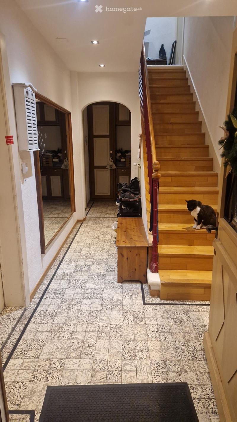 Spacious hallway with tiled flooring, wooden staircase leading upstairs, and a small bench with shoes placed on it. A cat is sitting on the stairs, and there is a mirror on the left wall.