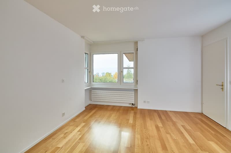 Empty room with light wooden flooring, white walls, and large windows offering a view of trees. A radiator is visible beneath the windows, and there is a door on the right side.