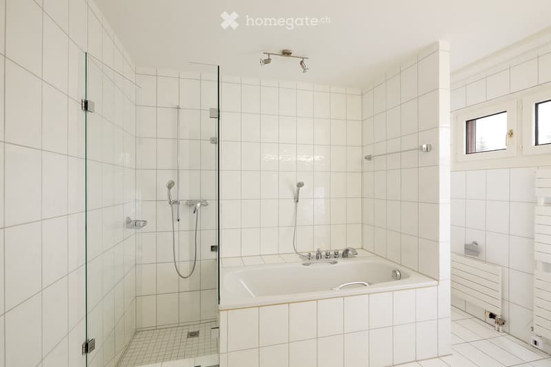 Modern bathroom with white tiled walls and floor, featuring a bathtub with a glass-enclosed shower area, multiple showerheads, and a towel rack.