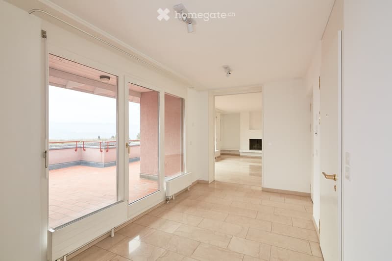 Empty hallway with tiled floor, white walls, large glass doors leading to a balcony with a view, and a visible fireplace in the adjacent room.