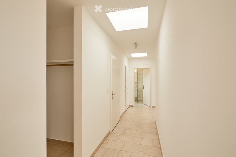 Clean, minimalist hallway with light-colored walls, tiled floor, and several doors leading to different rooms. Features a skylight and recessed lighting.