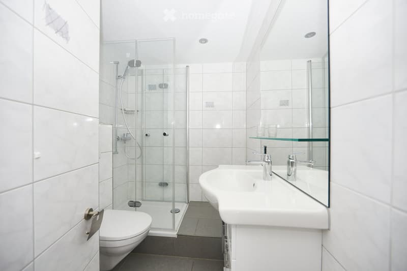 White tiled walls, glass shower enclosure, white sink, mirror, white toilet.