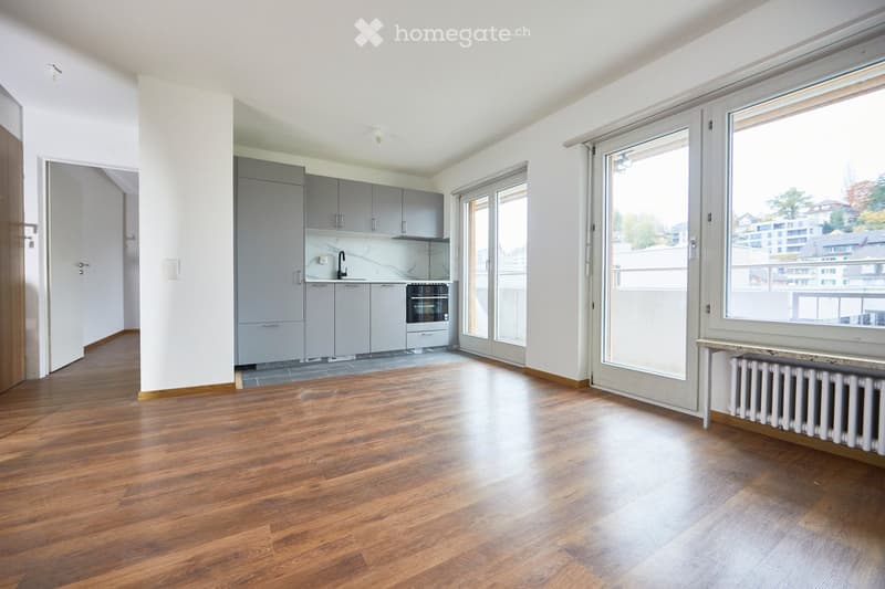 Spacious room, hardwood floors, white walls, large windows with balcony access, modern kitchen set up, built-in appliances