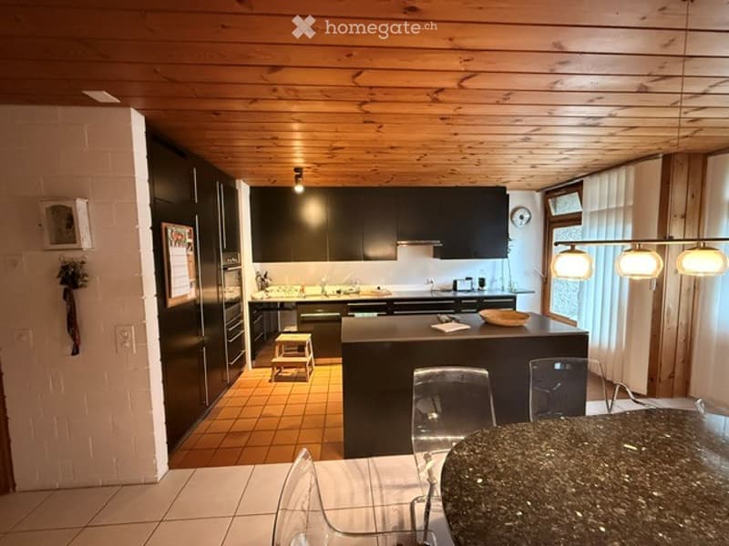 Modern kitchen with black cabinets, stainless steel appliances, and a central island with a granite countertop. The space includes a refrigerator, oven, sink, and a small wooden stool near the counter.