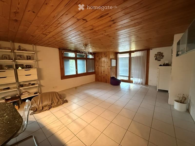 Bright, open room with wooden ceiling and walls, large windows, tiled floor, and minimal furnishings including a shelf, chair, and small table.