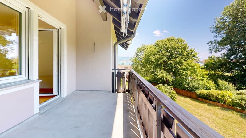 Balcony with wooden railing, open sliding door leading to interior, view of green garden and trees.