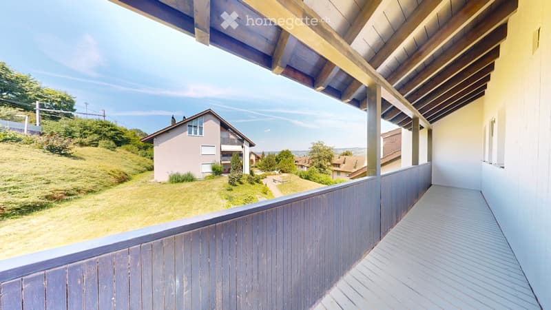 Balcony with wooden decking and railing, overlooking a grassy hillside with houses and trees in the background.