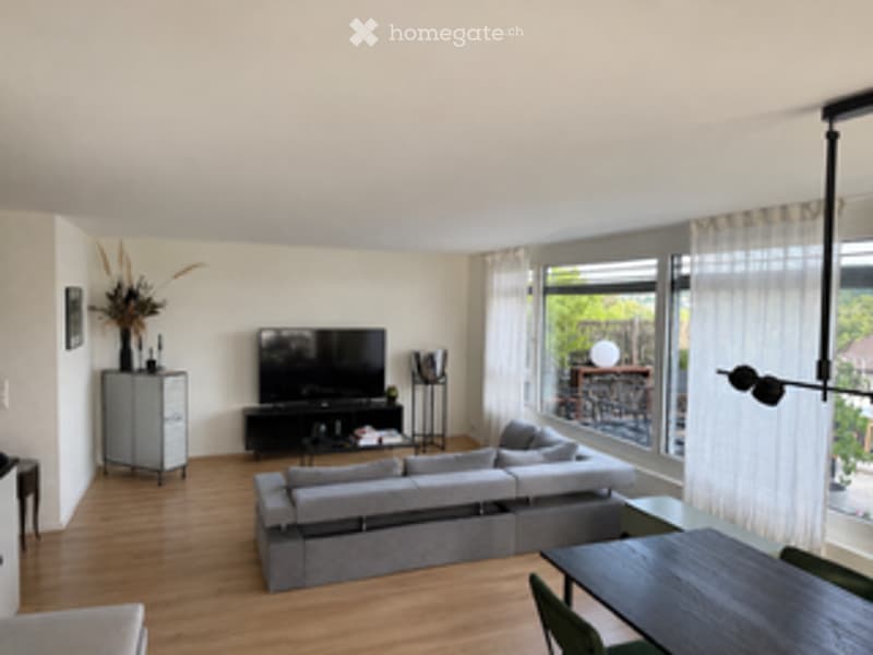 Modern living room with large windows and balcony access, featuring a grey sectional sofa, coffee table, TV stand, side table, dining table, and floor lamp.