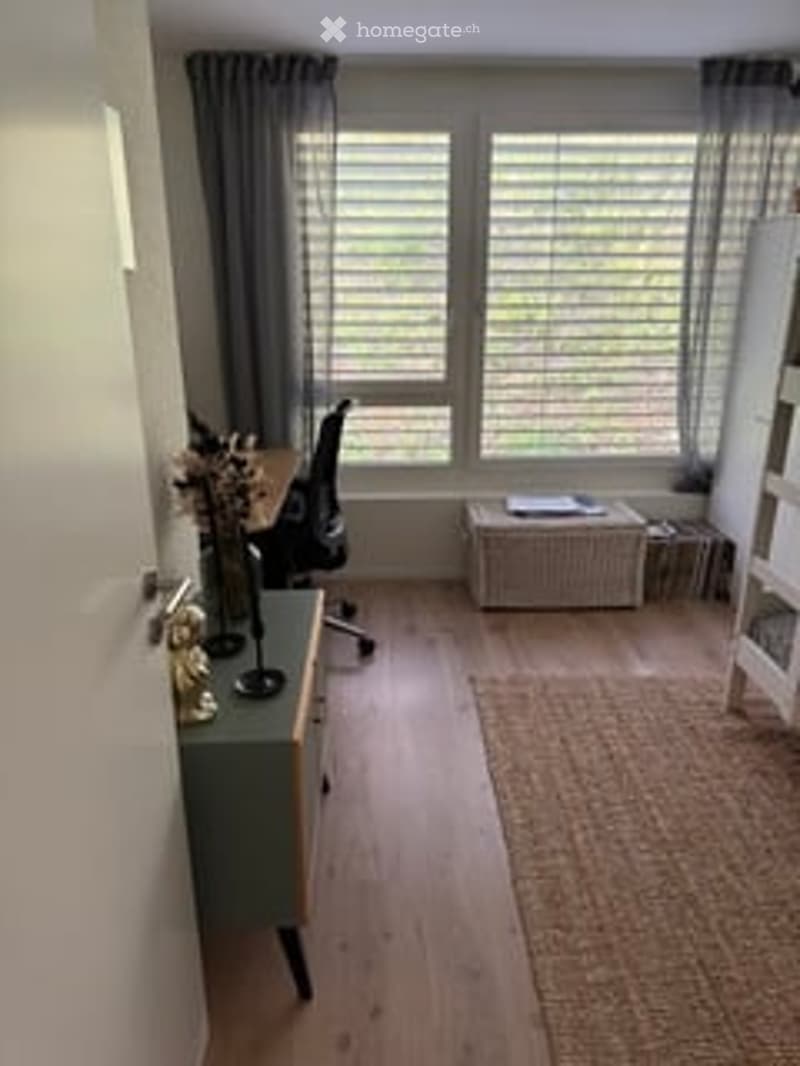 A modern, well-lit room with wooden flooring, large windows with blinds, a desk with a chair, a small cabinet, a vase on the cabinet, and a bookshelf with books.