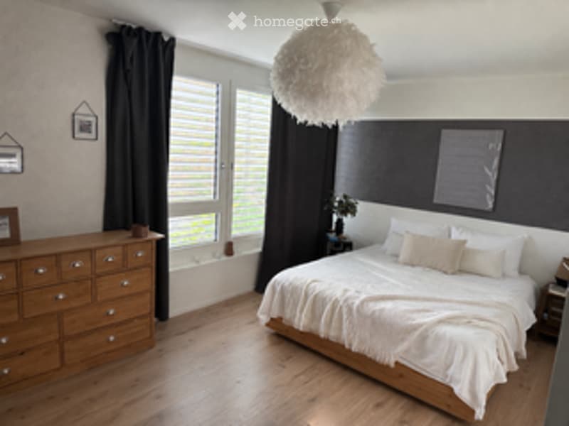 Modern bedroom with a large bed, nightstand, dresser, and large windows with dark curtains. The room features a wooden floor and a decorative hanging lamp.
