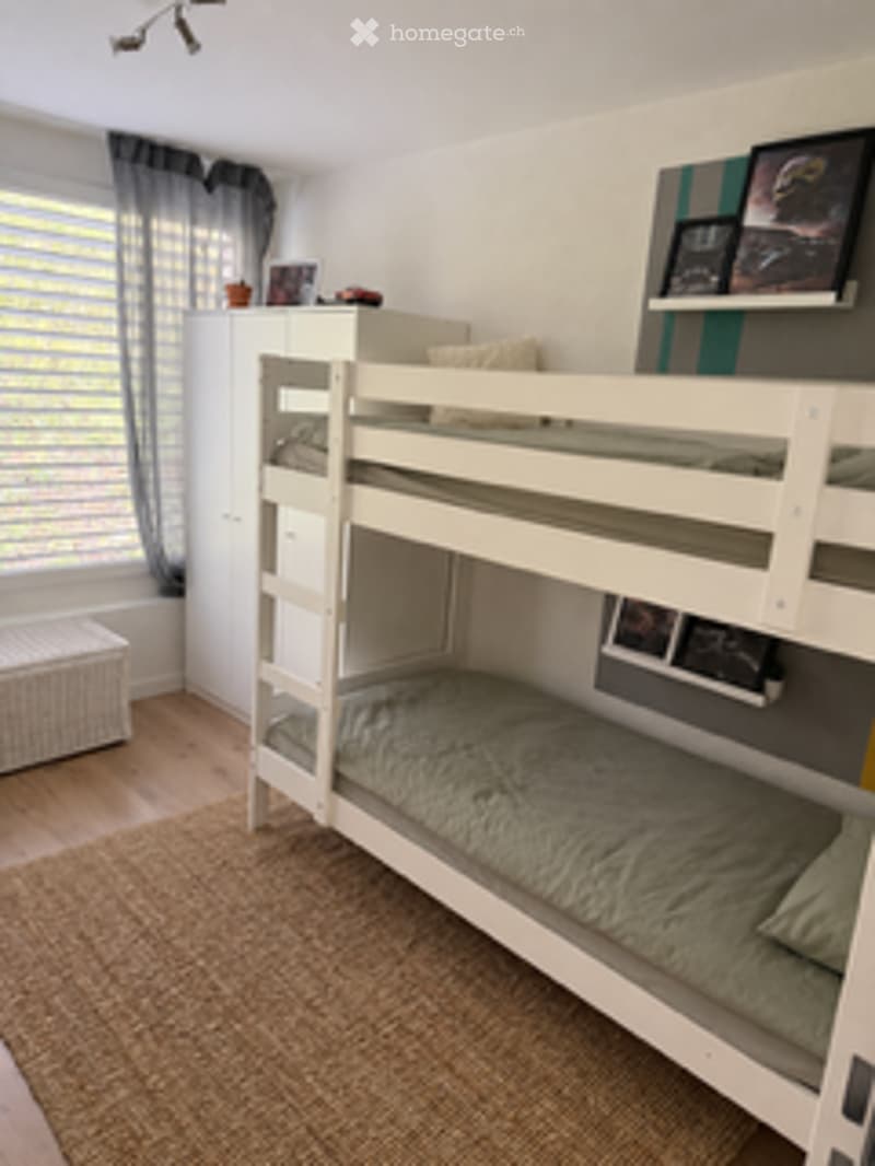 A cozy bedroom with a white bunk bed, pillows, and a blanket. The room features a nightstand, a curtain, and a picture frame on the wall.