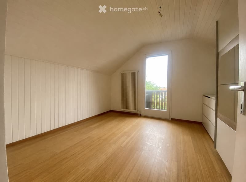 Empty room with wooden flooring, white walls, and a large window with a view of a balcony. The room features a radiator and a door with a modern handle.