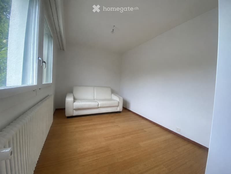 A modern living room with a white leather sofa, wooden flooring, and a radiator on the left side. Large windows allow natural light to fill the space.