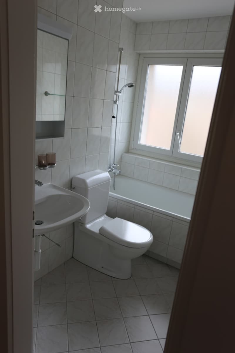 Modern bathroom with white tiles, a toilet, sink, bathtub, and shower. A small window provides natural light, and a mirror is mounted on the wall.