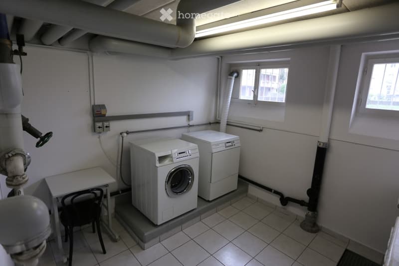 Utility room with tiled floor, two white washing machines, a small table, and a chair.
