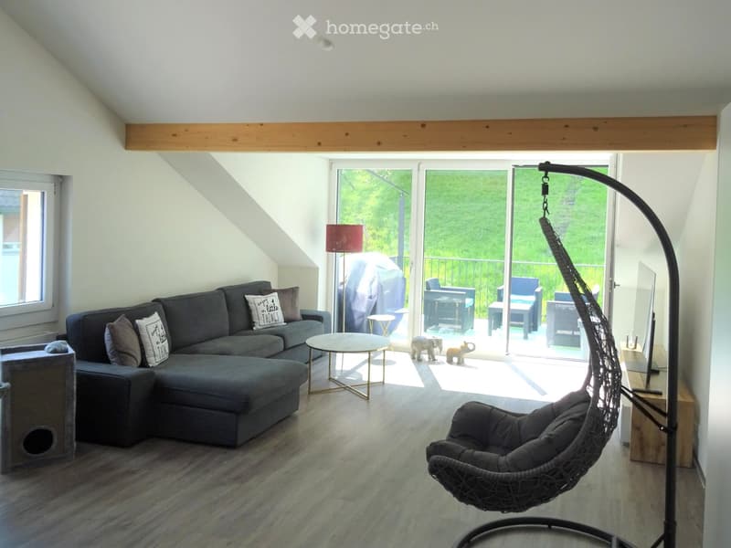 Modern living room with slanted ceiling, grey sectional sofa, wicker hanging chair, coffee table, floor lamp, and cat tree. Large sliding glass doors open to a balcony with outdoor furniture.